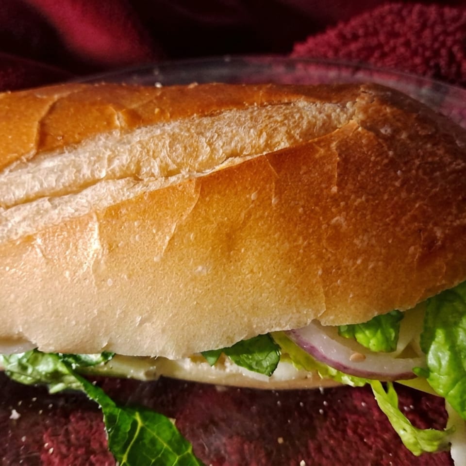 Crusty baguette sandwich with lettuce, red onion, and creamy filling on a glass plate over red fabric.