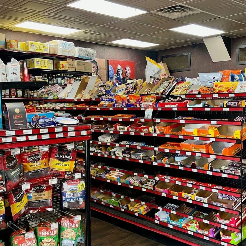 Convenience store shelves display snacks, candy, jerky, sauces, and packaged foods beneath fluorescent lighting.