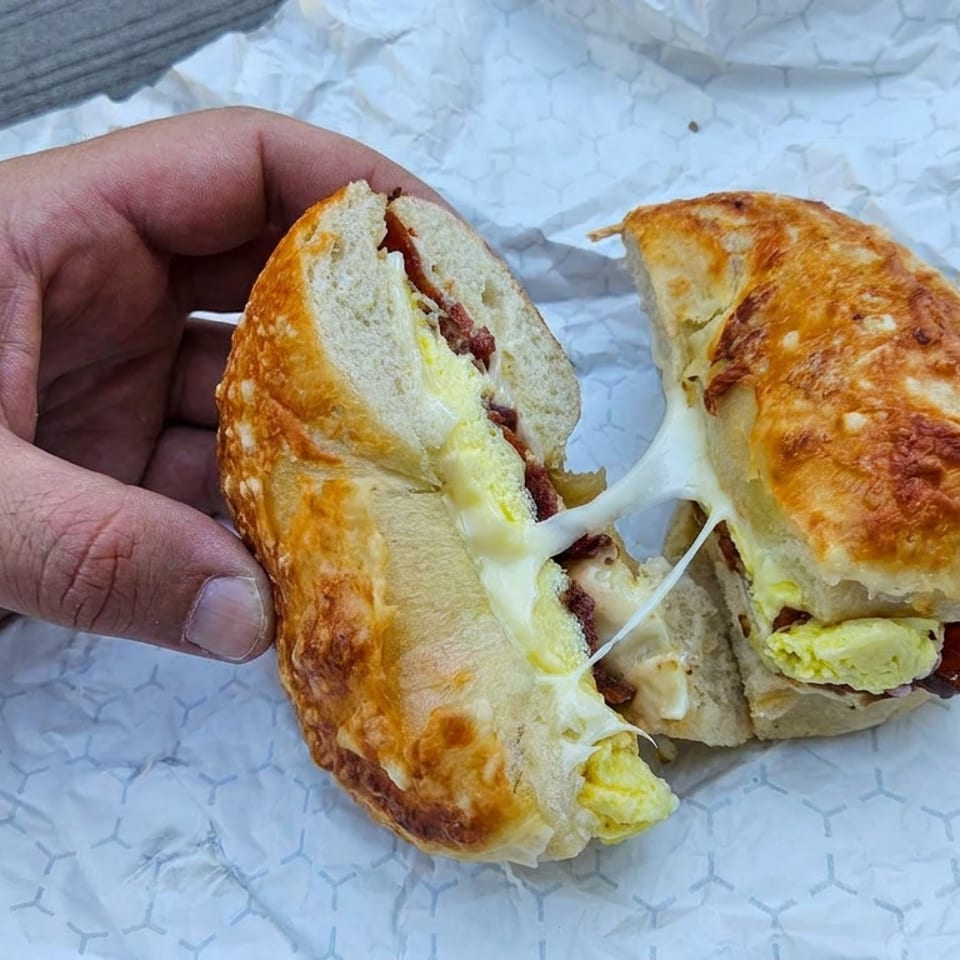Hand holds a toasted breakfast sandwich split open, showing melted cheese, egg, and bacon on patterned wrapping paper.