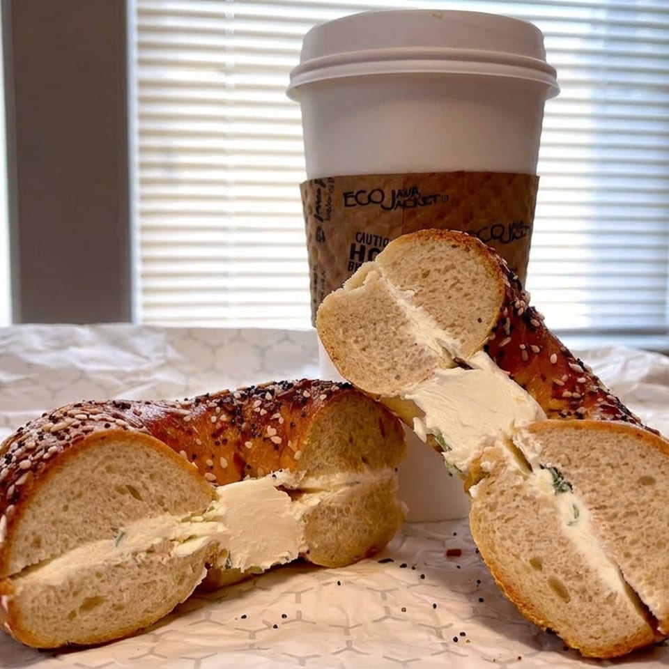 Sliced everything bagels spread with cream cheese sit on deli paper beside a takeaway coffee cup near sunlit windows.